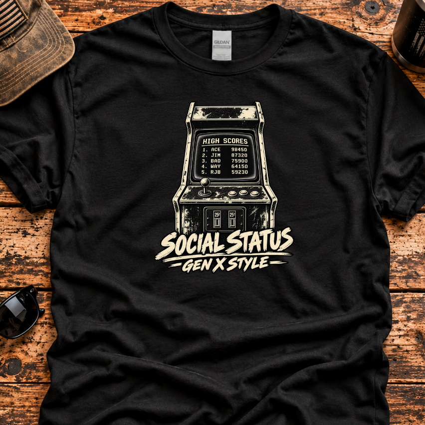 A black graphic t-shirt laid flat on a rustic wooden surface, featuring a vintage arcade machine illustration and the text 'SOCIAL STATUS GEN X STYLE'. The shirt is styled with a distressed cap, sunglasses, a tumbler, and a knife.