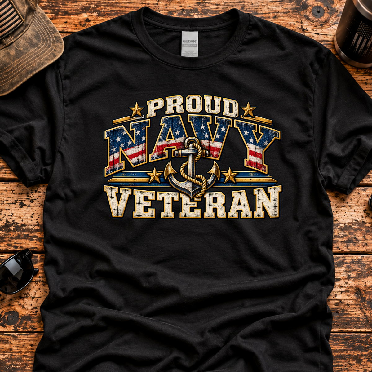 A black short-sleeve graphic T-shirt laid flat on a rustic wooden surface, featuring a distressed patriotic design that reads 'PROUD NAVY VETERAN' with an anchor and stars. A cap, sunglasses, coffee mug, are placed around it as styling props.