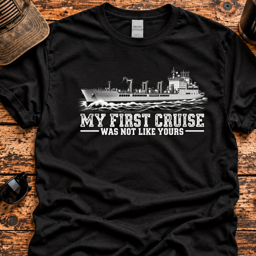 A black short-sleeve graphic T-shirt laid flat on a rustic wooden surface, with a white ship illustration and the text 'MY FIRST CRUISE WAS NOT LIKE YOURS' across the chest. A distressed cap, sunglasses, knife, and mug are placed around it as styling props.