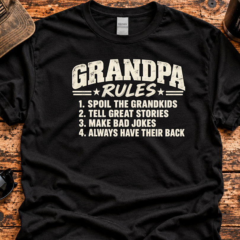 A black graphic t-shirt laid flat on a rustic wooden surface, featuring a white distressed print that reads 'GRANDPA RULES' followed by four numbered rules. The shirt is styled with a vintage cap, sunglasses, a coffee mug, and a knife around it.