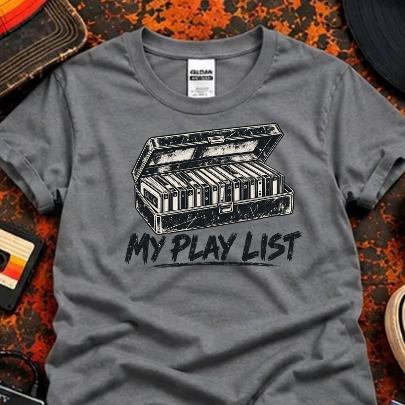 A heather gray graphic t-shirt featuring a black-and-white cassette tape illustration and the text 'MY PLAY LIST'. The shirt is styled in a flat-lay arrangement with retro props including a cassette tape, headphones, a vinyl record, jeans, and sneakers on a textured orange and black background.
