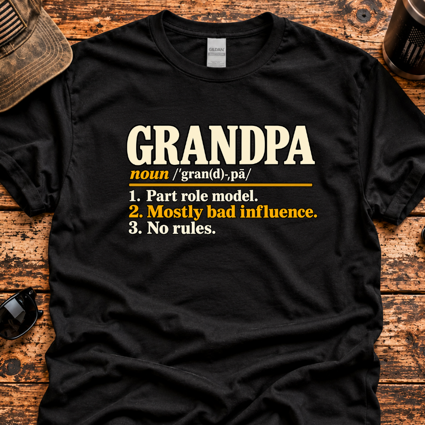 A black graphic t-shirt laid flat on a rustic wooden surface, featuring a humorous dictionary-style definition of 'GRANDPA' in cream and yellow text. The shirt is styled with a distressed brown cap, sunglasses,, and a coffee cup in the background.