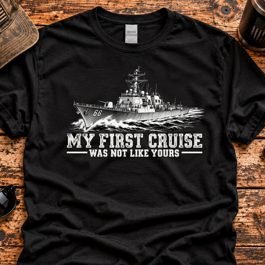 A black short-sleeve graphic T-shirt laid flat on a rustic wooden surface, featuring a white military ship illustration and the text 'MY FIRST CRUISE WAS NOT LIKE YOURS.' A distressed cap, sunglasses, coffee mug, and knife are placed around it as styling props.