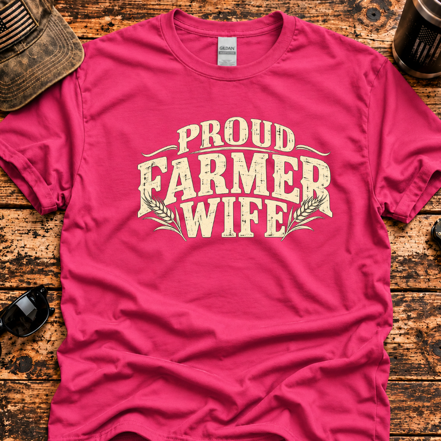 A bright pink t-shirt with a distressed cream-colored graphic reading 'PROUD FARMER WIFE' laid flat on a rustic wooden table, surrounded by lifestyle props including a cap, sunglasses, a coffee mug.