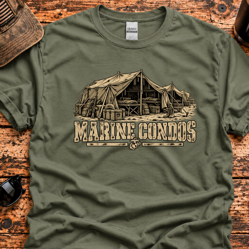 An olive green crewneck t-shirt with a vintage-style tan graphic of a military tent and the text 'MARINE CONDOS'. The shirt is laid flat on a rustic wooden table surrounded by lifestyle props including a baseball cap, sunglasses, a coffee mug, and a knife.