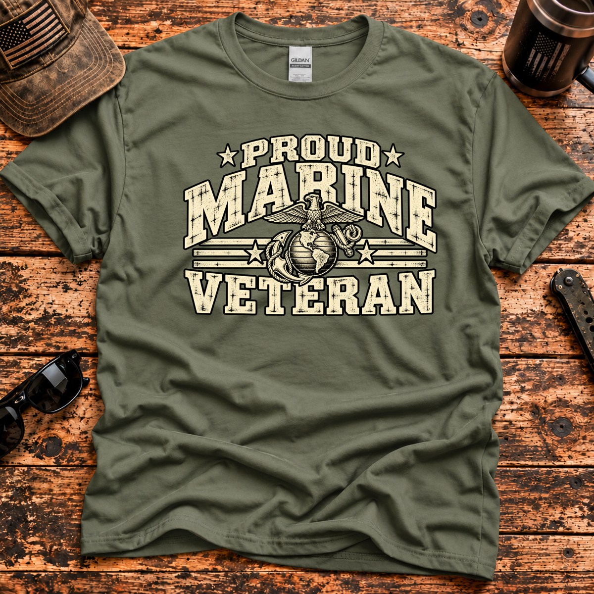 An olive green t-shirt with a distressed cream graphic reading 'PROUD MARINE VETERAN' and featuring the Marine Corps emblem, laid flat on a rustic wooden background with lifestyle props.