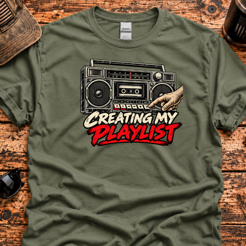 An olive green graphic t-shirt laid flat on a rustic wooden surface, featuring a retro boombox and a hand pointing with the text 'CREATING MY PLAYLIST' in white and red. Lifestyle props like a baseball cap, sunglasses, a knife, and a coffee cup are scattered around it.