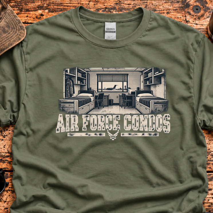 An olive green graphic t-shirt laid flat on a rustic wooden table, surrounded by lifestyle props including a baseball cap, sunglasses, a coffee mug, and a knife. The t-shirt features a black-and-white graphic of an Air Force living room with the text 'AIR FORCE CONDOS' and a winged emblem.