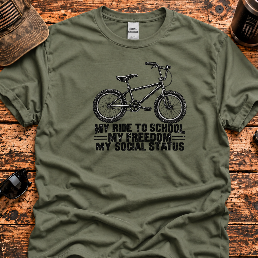 An olive green short-sleeve t-shirt with a black graphic of a BMX bicycle and the text 'MY RIDE TO SCHOOL / MY FREEDOM / MY SOCIAL STATUS', laid flat on a rustic wooden surface with lifestyle props like a cap, sunglasses, and a coffee cup.