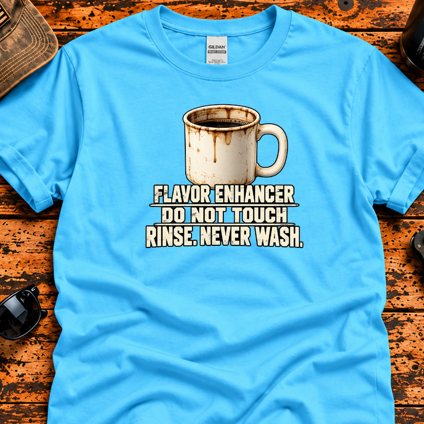 A bright blue graphic t-shirt laid flat on a rustic wooden surface, surrounded by lifestyle props including a cap, sunglasses, a folding knife, and a tumbler. The shirt features a graphic of a dripping coffee mug with the text 'FLAVOR ENHANCER DO NOT TOUCH RINSE.