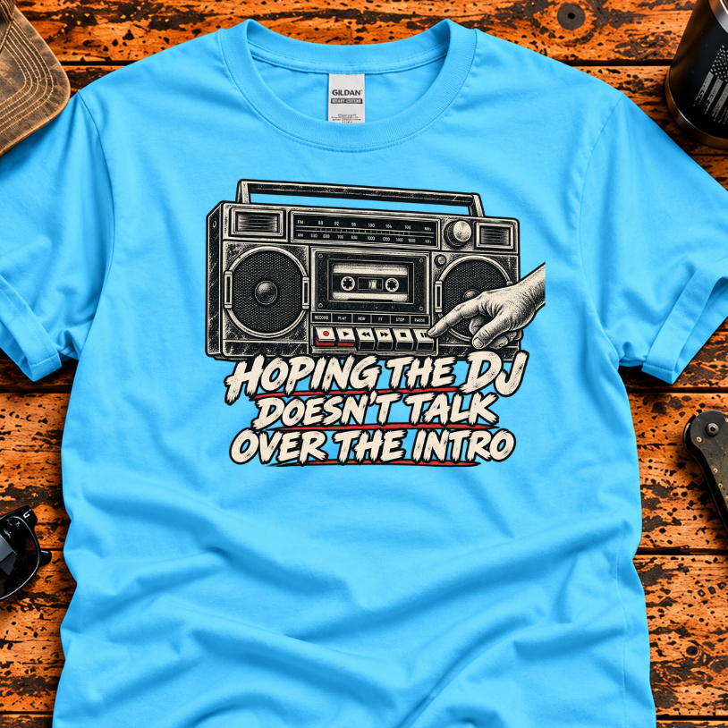 A bright blue graphic t-shirt laid flat on a rustic orange-brown wooden surface, surrounded by lifestyle props including sunglasses, a folding knife, a cap, and a tumbler. The t-shirt features a black-and-white retro boombox graphic with a hand adjusting the volume and the text 'HOPING THE DJ DOESN'T TALK OVER THE INTRO'.