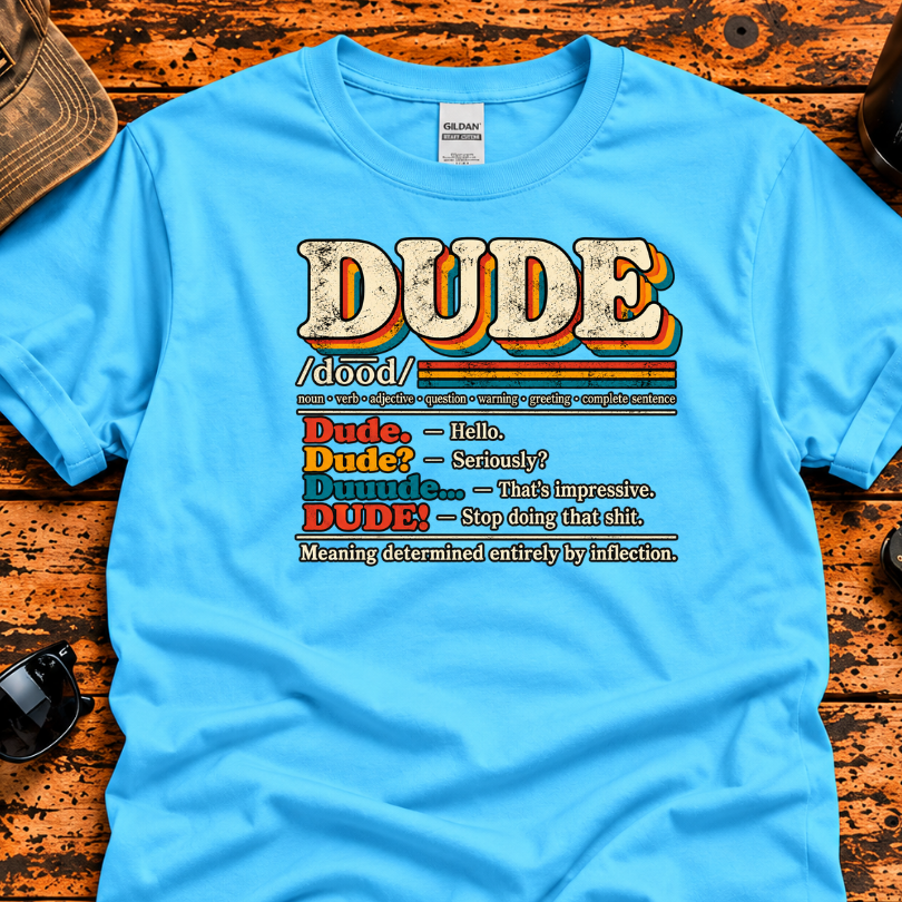 A bright blue graphic t-shirt laid flat on a rustic wooden surface, surrounded by lifestyle props including a baseball cap, sunglasses,, and a drink can. The t-shirt features a retro-style dictionary definition graphic for the word 'DUDE' with humorous dictionary-style entries.