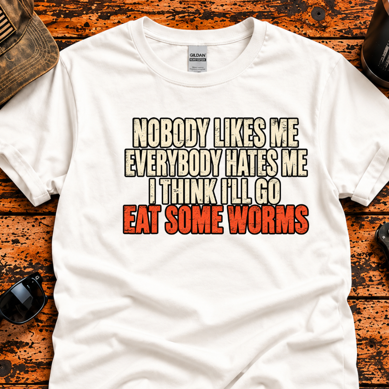 A white graphic t-shirt laid flat on a rustic wooden background with various outdoor-style props including a camouflage cap, sunglasses, a folding knife, and a tumbler. The t-shirt features a bold text graphic that reads 'NOBODY LIKES ME EVERYBODY HATES ME I THINK I'LL GO EAT SOME WORMS' in beige, black, and red lettering.
