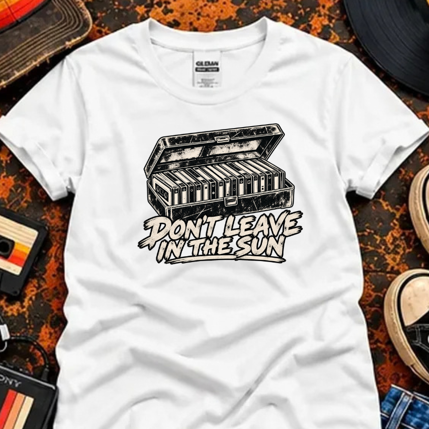 A white short-sleeve graphic T-shirt laid flat on a textured orange-brown surface with vintage music props around it. The shirt has a black illustrated cassette deck graphic and the phrase “DON’T LEAVE IN THE SUN” across the chest.