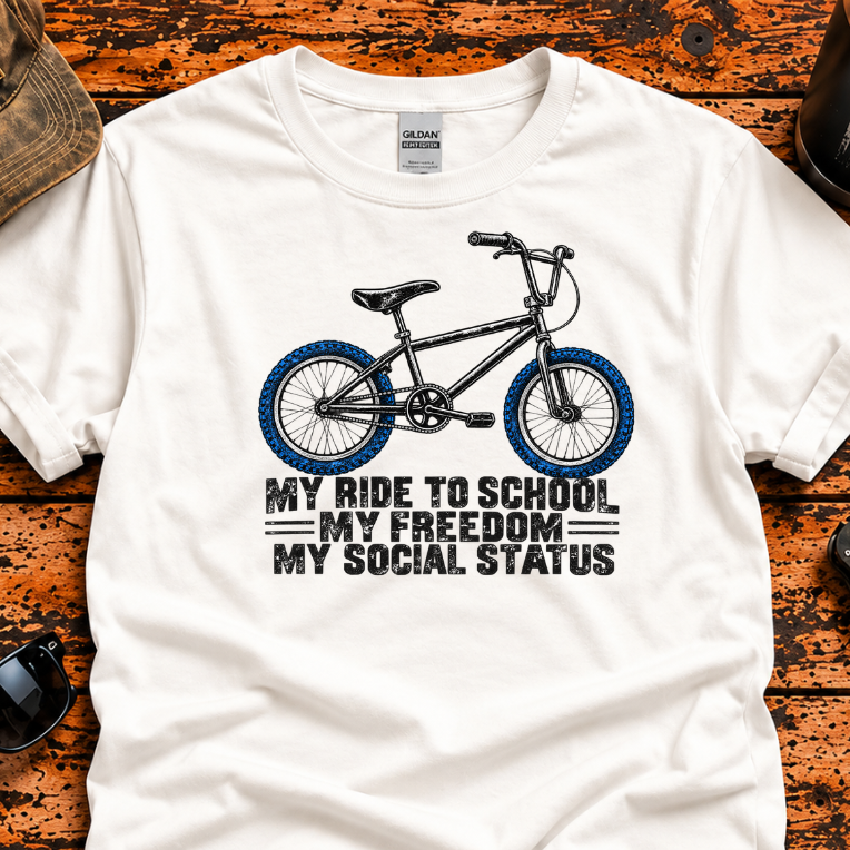 A white short-sleeve graphic T-shirt laid flat on a rustic orange-brown wooden surface, surrounded by lifestyle props including a cap, sunglasses, a tumbler, and a folding knife. The shirt features a black bicycle graphic with blue wheels and the text 'MY RIDE TO SCHOOL MY FREEDOM MY SOCIAL STATUS'.