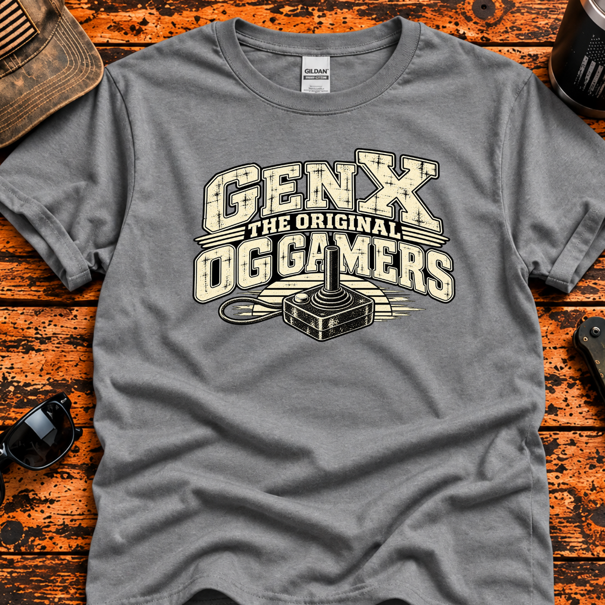 A heather gray graphic t-shirt laid flat on a distressed orange-brown wooden surface, surrounded by lifestyle props including a cap, sunglasses, a tumbler. The t-shirt features a distressed cream and black graphic that reads 'GENX THE ORIGINAL OGGAMERS' with a retro computer mouse illustration.