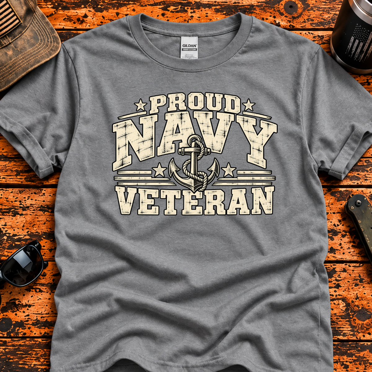 A heather gray t-shirt with a distressed cream and black graphic reading 'PROUD NAVY VETERAN' with an anchor design, laid flat on a rustic orange-brown wooden surface with lifestyle props including a cap, sunglasses, a tumbler.