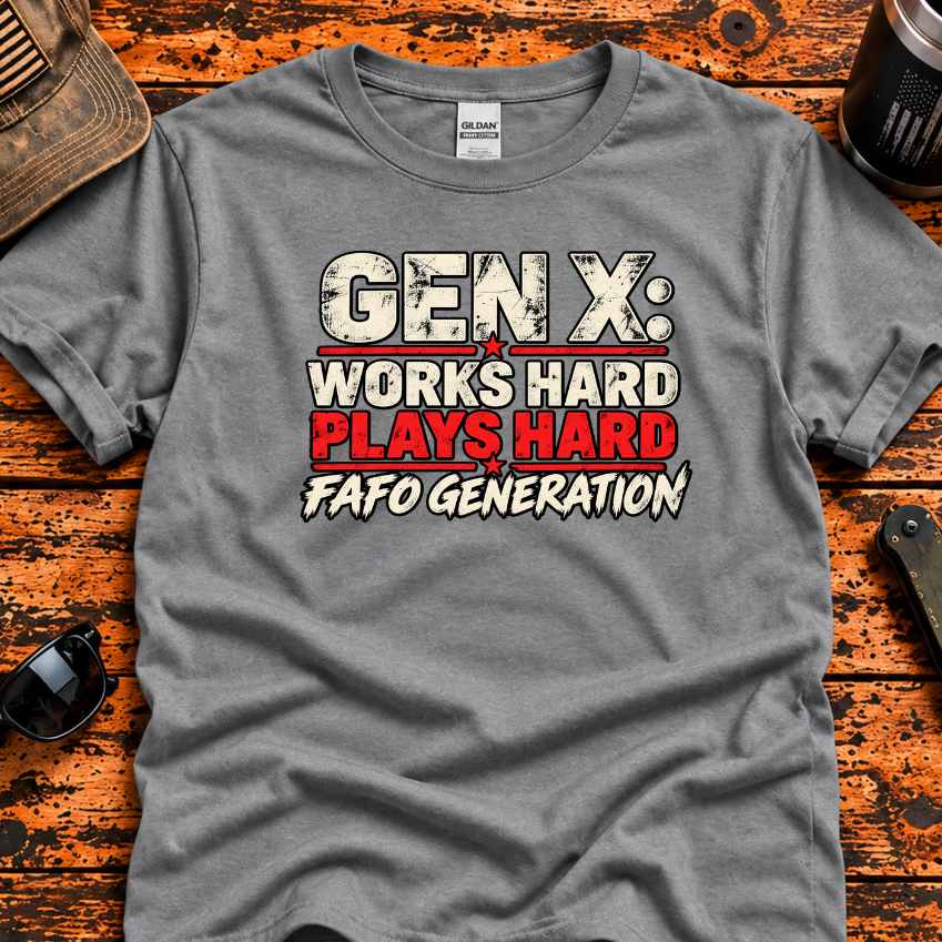 A heather gray graphic t-shirt laid flat on a rustic orange-brown wooden surface, surrounded by lifestyle props including a cap with an American flag, sunglasses, a tumbler, and a folding knife. The t-shirt features a distressed graphic that reads 'GEN X: WORKS HARDPLAYS HARD FAFO GENERATION'.