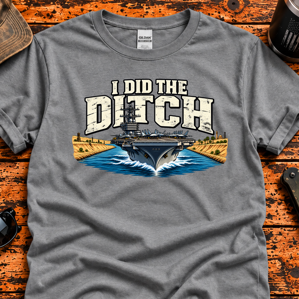 A heather gray graphic t-shirt laid flat on a rustic orange-brown wooden table, surrounded by lifestyle props including sunglasses, a tumbler, a hat. The t-shirt features a graphic of a military aircraft carrier on blue water with the text 'I DID THE DITCH' in distressed white letters.