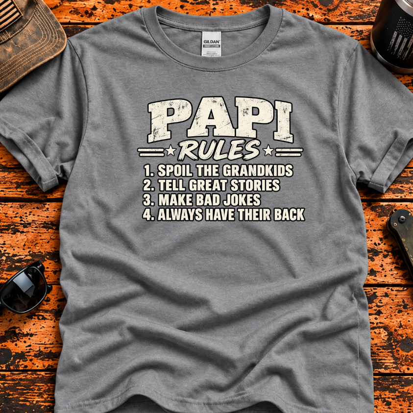 A heather gray graphic t-shirt laid flat on a rustic wooden surface, surrounded by lifestyle props including sunglasses, a folding knife, a tumbler, and a distressed cap. The t-shirt features a white graphic print that reads 'PAPI RULES' followed by a numbered list of four humorous rules.