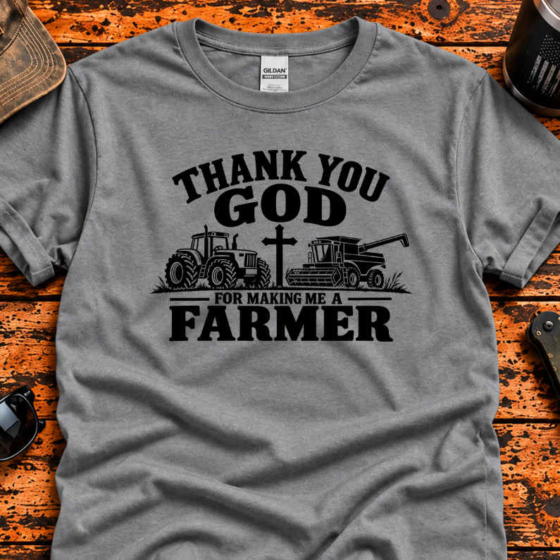 A heather gray short-sleeve t-shirt with a black graphic print that reads 'THANK YOU GOD FOR MAKING ME A FARMER' with illustrations of a tractor, a cross, and a combine harvester. The shirt is displayed in a flat-lay arrangement on a rustic, weathered orange-brown wooden table, surrounded by lifestyle props including sunglasses, a tumbler, and a distressed baseball cap.