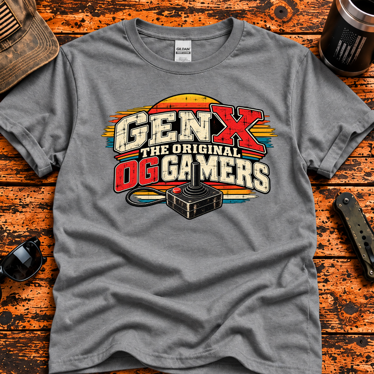 A heather gray graphic t-shirt laid flat on a rustic orange-brown wooden surface, surrounded by lifestyle props including a baseball cap, sunglasses, a tumbler. The t-shirt features a retro-style graphic that reads 'GEN X THE ORIGINAL DG GAMERS' with a vintage computer mouse illustration.