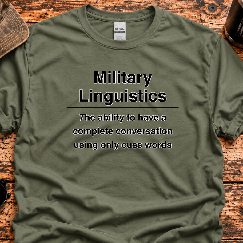 An olive green crewneck t-shirt with black printed text reading 'Military Linguistics' and a humorous definition, laid flat on a rustic wooden surface with lifestyle props including a cap, sunglasses, and a coffee cup.